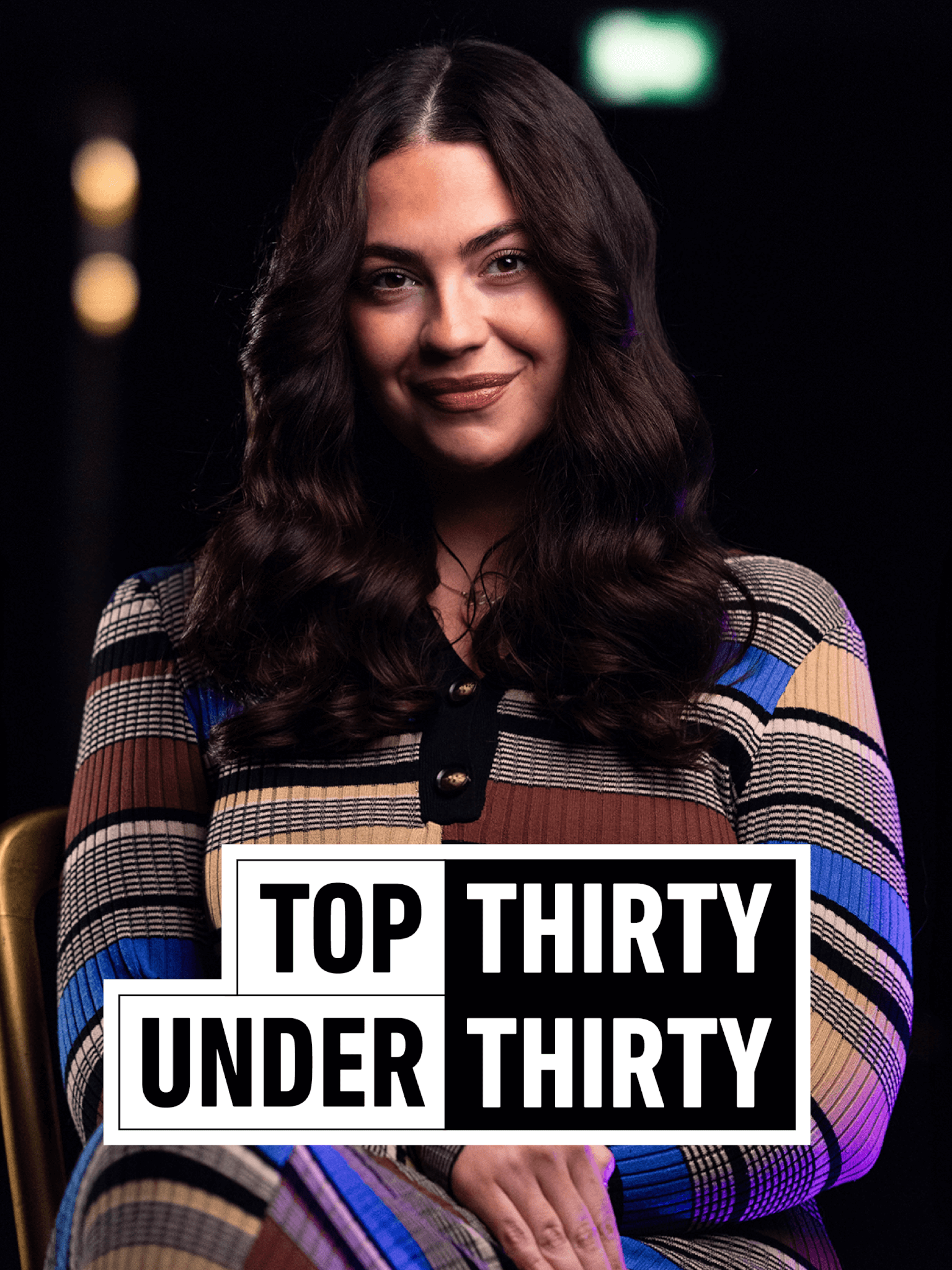 Creative Lead, Rachel Smith sit centre shot in a colourful dress, a black background with blurred spotlights sits behind her. The text logo in front of her reads: TOP THIRTY UNDER THIRTY.