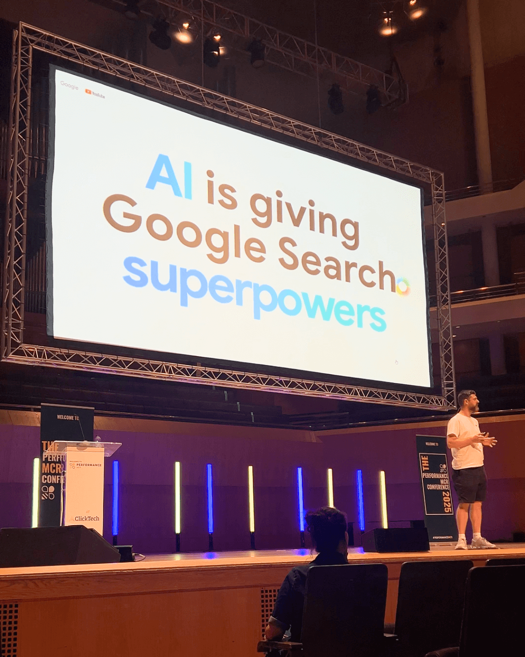 The image shows a marketing expert delivering a talk on AI. The words on screen behind him rad: "AI is giving Google Search superpowers."